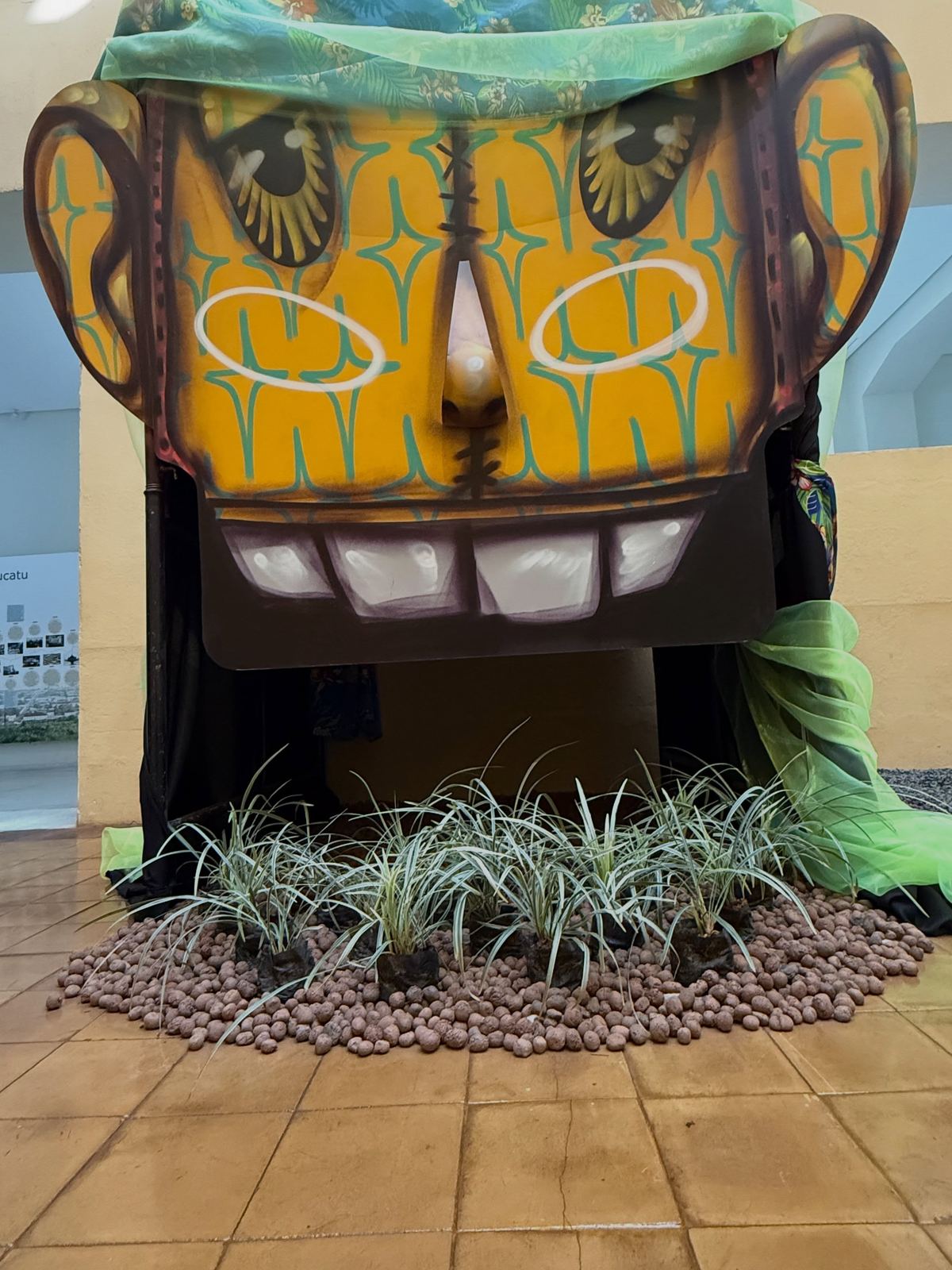 Escultura de rosto amarelo sobre plantas na exposição A Casa