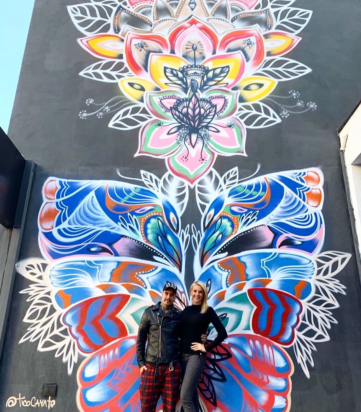 Mural de mandala e borboleta numa empena, com o artista na base