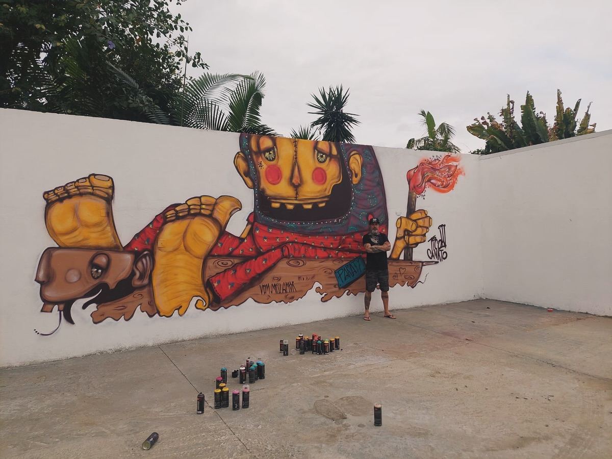 Mural de grande escala num muro branco, com o artista pequeno ao pé da obra e latas de spray no chão
